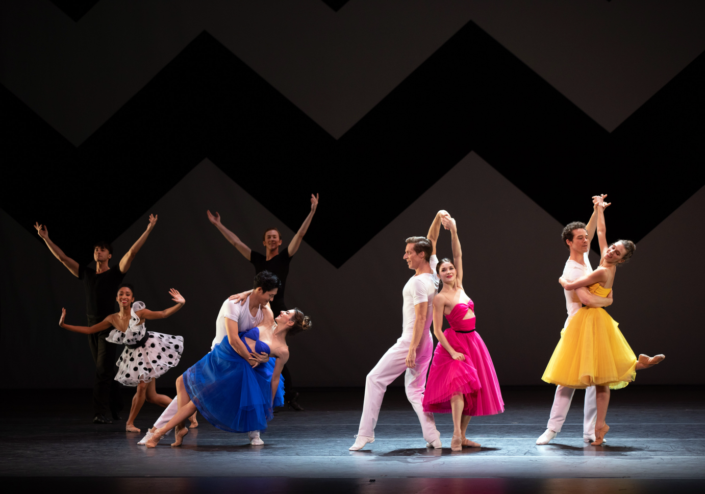 Three couples pose in a line with classic dips and closed position dance holds. On the left side, two men extend their arms in a V while a woman in a polka-dot dress places her arms to the side, wrists flexed.